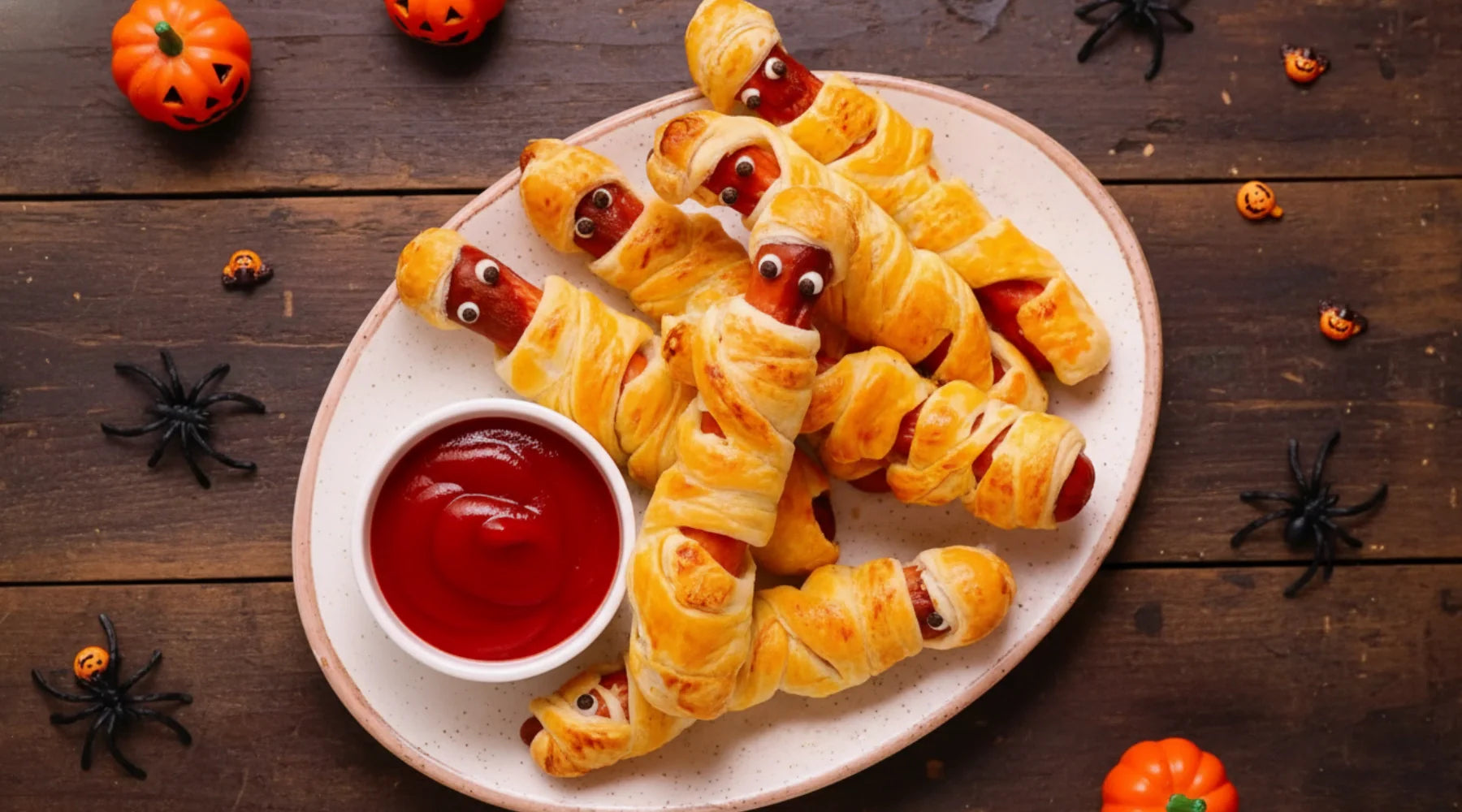 Hot-dogs momies pour Halloween — saucisses enroulées dans de la pâte feuilletée, décorées avec des yeux comestibles et servies avec du ketchup, sur un fond festif avec des citrouilles et araignées décoratives.